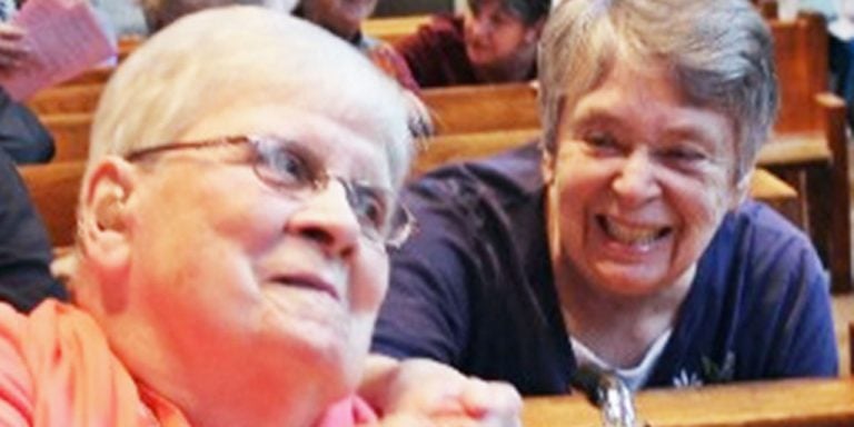 Two women pictured candidly, one woman is sitting and smiling, leaning forward placing her hand on the shoulder of her friend sitting in front of her. The friend in front is smiling and tilting her head to hear what her friend behind her has to say.