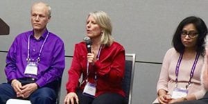 One man and two women sit together on a stage at an event. They are all three wearing matching lanyards and the woman sitting in the middle is speaking into a microphone.