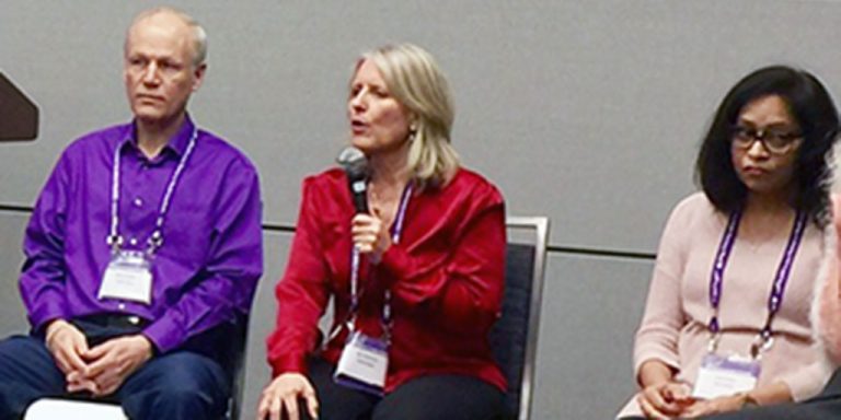 One man and two women sit together on a stage at an event. They are all three wearing matching lanyards and the woman sitting in the middle is speaking into a microphone.