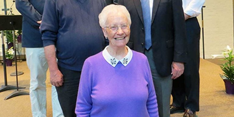 Six men wearing dark grey and black suits standing on a higher platform indoors and smiling with one woman standing lower, in the middle wearing a bright purple sweater.