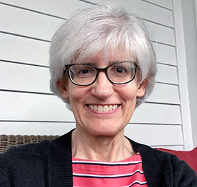A woman with short white hair and black-framed glasses smiling brightly for a headshot picture wearing a pink striped blouse and a black cardigan.