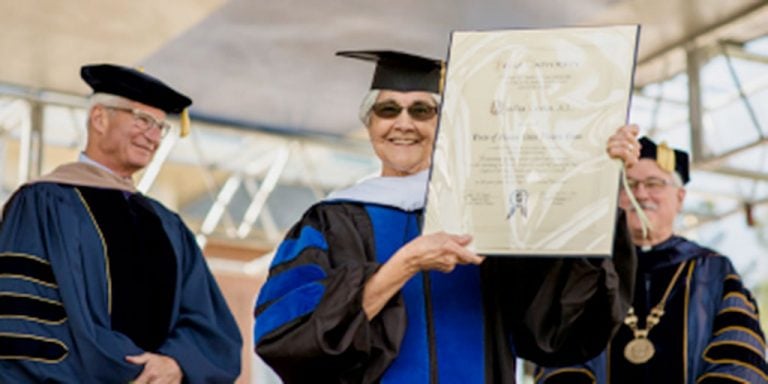 A woman in ceremonial graduation attire smiling on stage displaying a large white plaque award with two men in ceremonial graduation attire behind her.