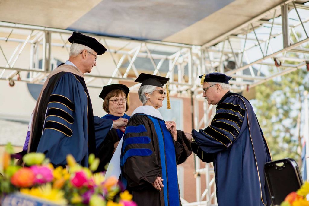 The university president and two others in ceremonial robes assists the graduate with her doctoral academic dress on stage outdoors.