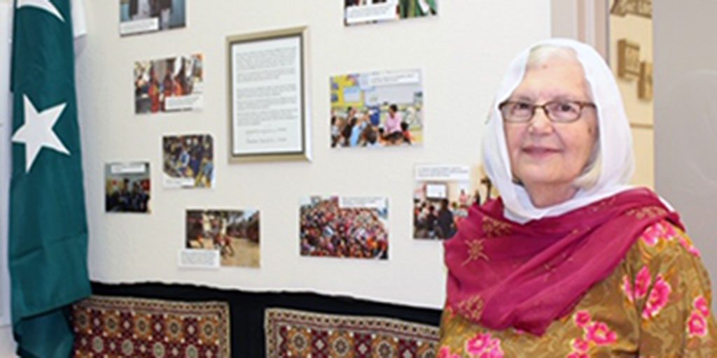 A woman wearing Pakistani attire smiling brightly in a classroom next to a collage wall of Pakistani appreciation.