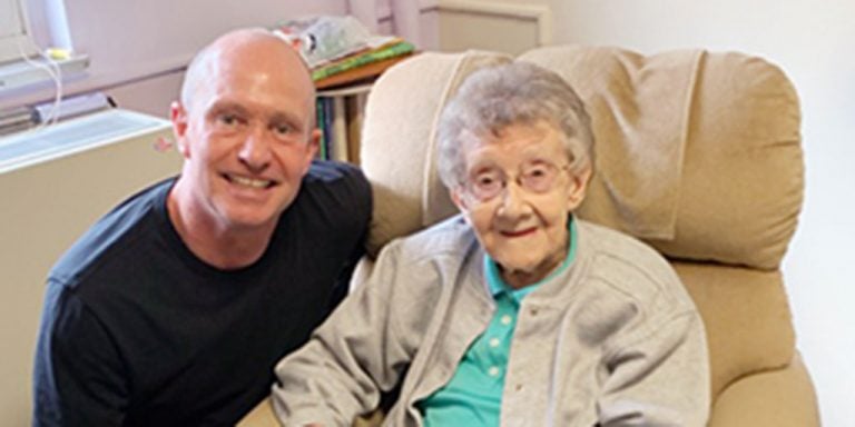 A woman with short grey hair and wire-framed glasses smiling while sitting in a beige recliner chair with a bald man crouched next to her smiling for the picture with her.