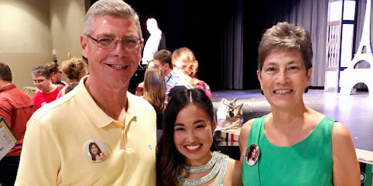 Two women and a man smiling together for a group picture at a social event. The women are wearing dresses, the man is wearing a collared shirt.