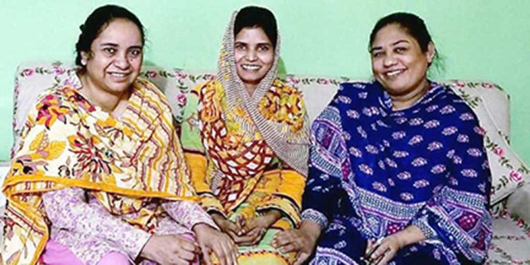 Three women wearing colorful patterned head scarves smiling together while sitting on a couch in a room with light green walls.