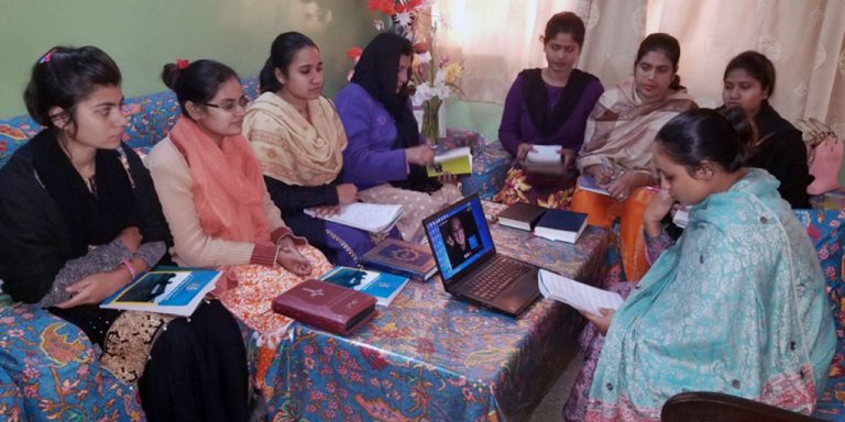 8 women sitting together in a living room on three couches in a u formation. They are wearing colorful cultural clothing, and some are wearing head scarves.