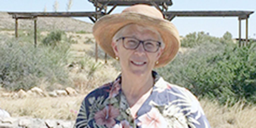 A woman wearing a sun hat, black-framed glasses, and a tropical patterned button-up shirt smiling for a picture outdoors in the desert on a sunny day.