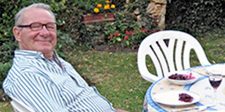 Two men sitting across from each other at a round table outdoors in a grass yard enjoying a meal. There are other empty chairs around them with additional plates of food and drinks.