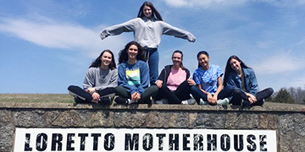 Five girls sitting and one girl standing and posing with her arms out to the side smiling together for a group picture on top of a stone sign for Loretto Motherhouse on a sunny day.
