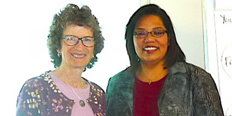 Two women standing together and smiling for a picture together in a bright room.