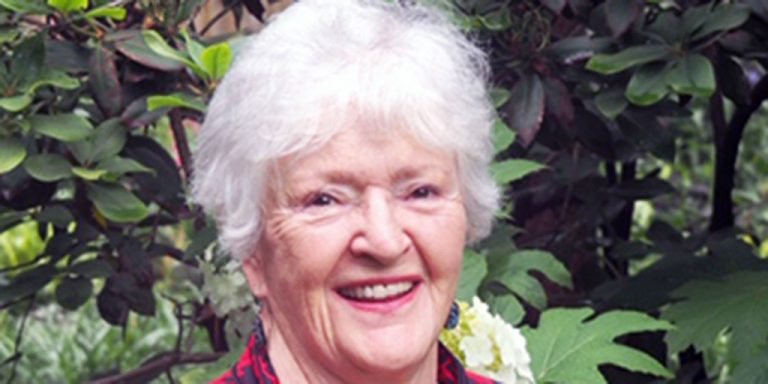 A woman with short white hair wearing a black and red patterned blouse smiling for a portrait picture with a lush greenery background.