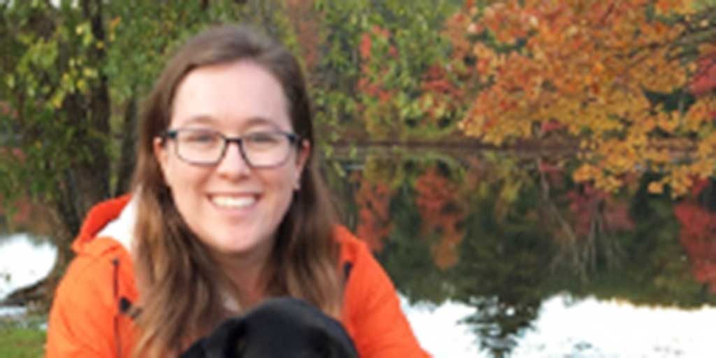 A woman, Erica Stewart, with long brown hair, black-framed glasses, and a bright orange rain coat smiling brightly while crouching down next to her black lab dog on a walk in front of a big body of water on a gloomy autumn day.