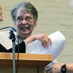 Three women conversing at a wooden podium with a microphone indoors in front of a white brick wall. They are exchanging papers in hand too.