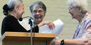 Three women conversing at a wooden podium with a microphone indoors in front of a white brick wall. They are exchanging papers in hand too.