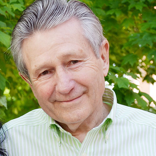 A man with short, grey hair smiling brightly for a headshot picture in front of greenery. He is wearing a light green and white plaid collared shirt.