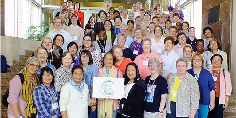 A large group photo on the front stairs of a building. The woman in the center of the first row is holding a poster board with the conference logo.