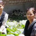 Two women smiling while preoccupied with harvesting leafy greens in an outdoor garden on a sunny day.