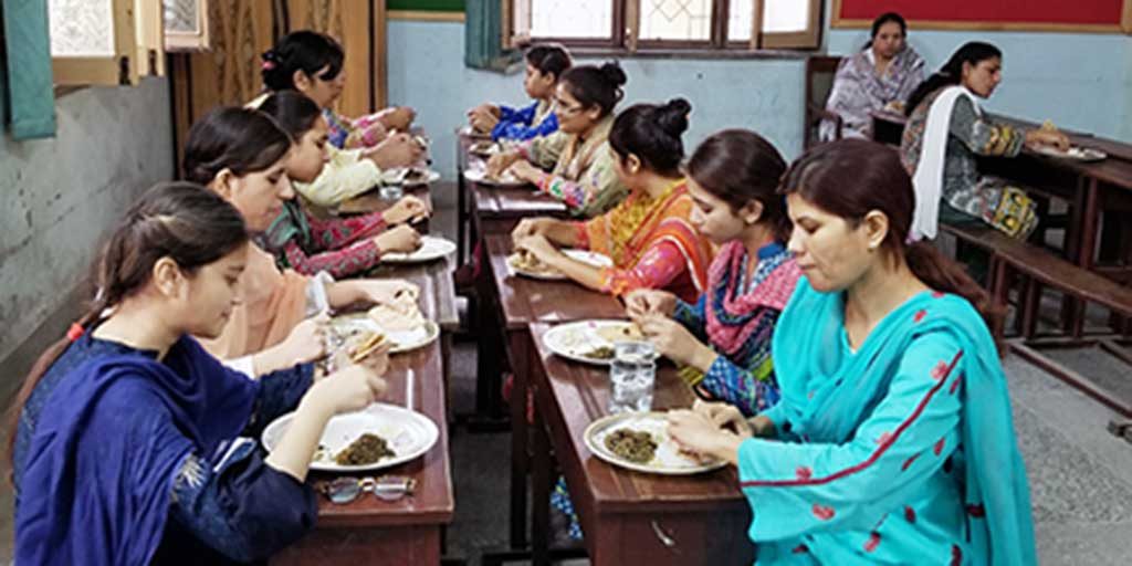 Nine women sitting together enjoying a meal at cafeteria-style benches and tables indoors on a sunny day.