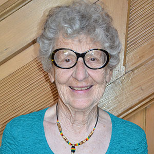 A woman with short, grey hair smiling warmly wearing a bright aqua blue shirt, beaded necklace and big black circular framed glasses. She is standing in front of a plain wooden wall.