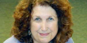 A woman, Paulette Peterson, with long curly brown hair and big earrings wearing a blue collared shirt smiling brightly for a headshot picture with a plain green background.