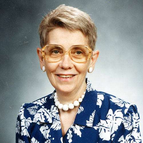 A woman with short, light brown hair and big round glasses, smiling for a portrait picture with a grey vignette background while wearing a navy blue and white patterned blouse and a white necklace with matching earrings.