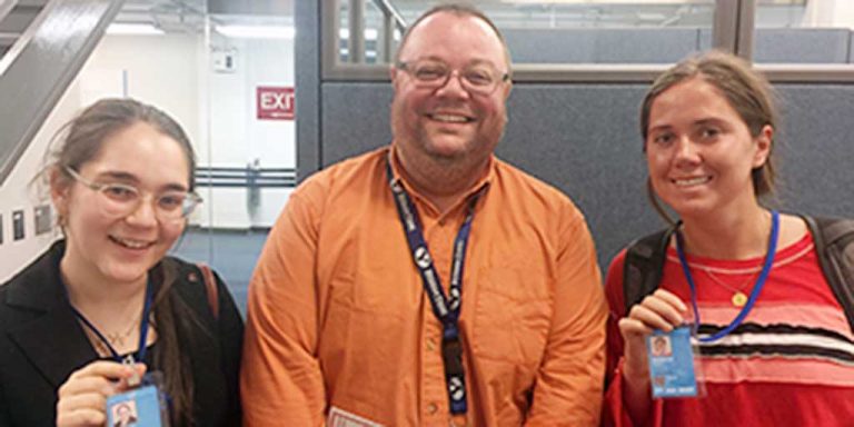 Two women with long brown hair pulled back and a man standing in the middle smiling together for a group picture indoors. They are all wearing matching lanyards with blue cards on the end.