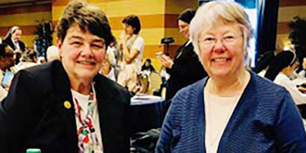 Two women, Cathy Mueller and Claire Skyes, smiling brightly together for a picture indoors aat a social event. They are both wearing dress clothes, one woman has short brown hair and the other has short grey hair and glasses.