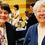 Two women, Cathy Mueller and Claire Skyes, smiling brightly together for a picture indoors aat a social event. They are both wearing dress clothes, one woman has short brown hair and the other has short grey hair and glasses.