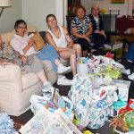 Nine women sitting together in a living room with assorted donations. They are sorting, and planning for donations to be given to those in need.