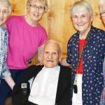 Four women standing and smiling surrounding a man in a wheelchair for a group picture. They are all wearing dress clothes, it is the man in the center's 90th birthday.