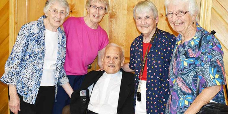 Four women standing and smiling surrounding a man in a wheelchair for a group picture. They are all wearing dress clothes, it is the man in the center's 90th birthday.