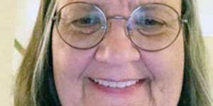 A woman, Jane Wilson with shoulder-length light-colored hair and round wire-framed glasses smiling brightly for a selfie picture indoors.
