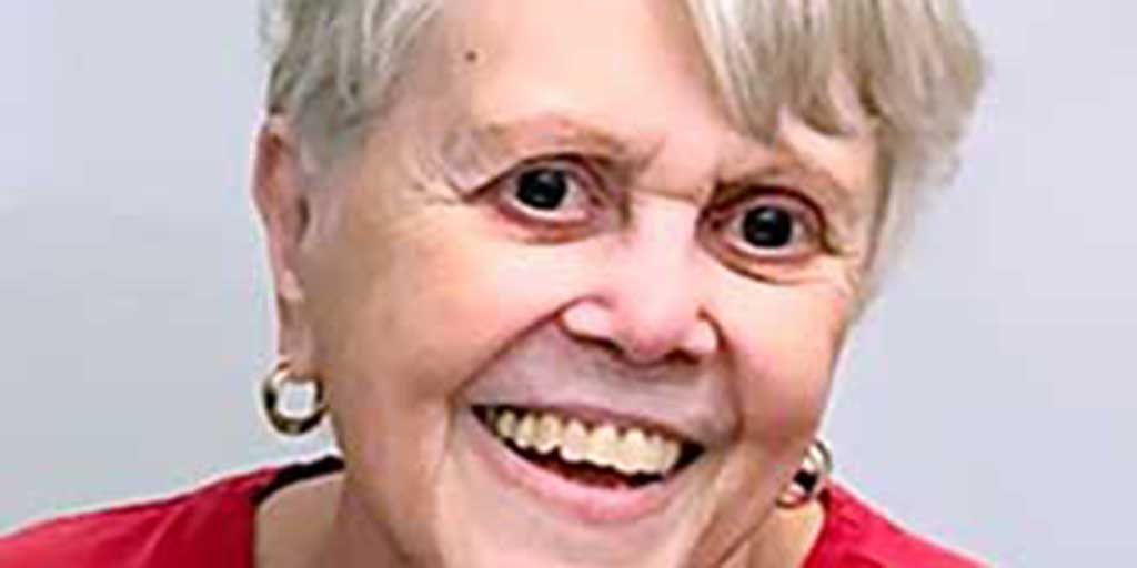 A woman, Lillian Moskeland, with shirt white hair, gold hoop earrings, and a red shirt smiling brightly for a portrait picture with a plain white background.