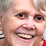 A woman, Lillian Moskeland, with shirt white hair, gold hoop earrings, and a red shirt smiling brightly for a portrait picture with a plain white background.
