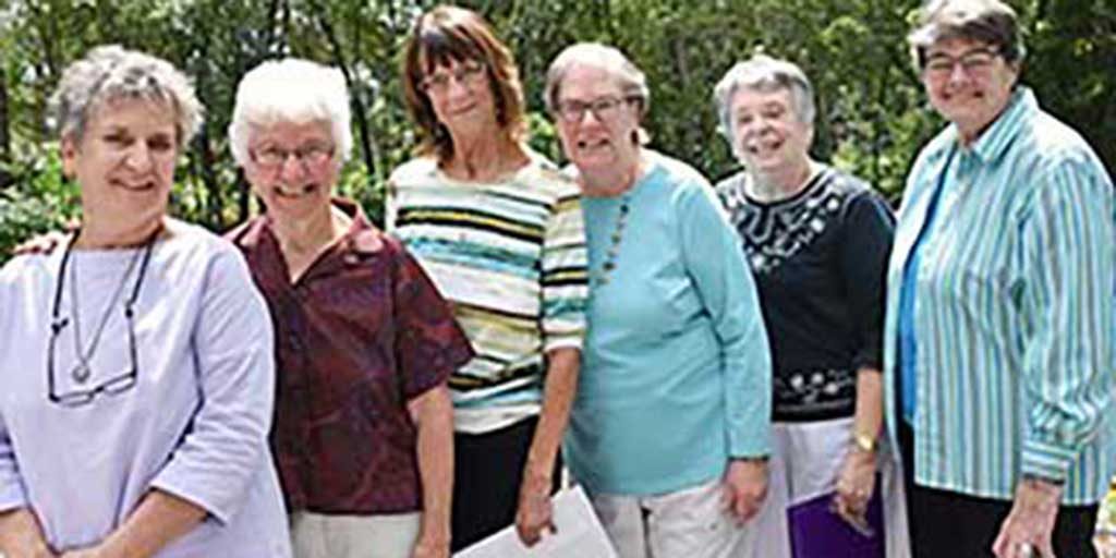 Six women smiling together for a group picture outdoors on a sunny day in front of large leafy green trees.