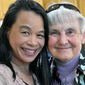Two women smiling brightly for a picture together indoors with their heads touching.