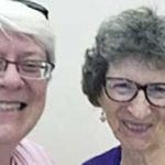 Two women, Beth Blissman and Jean East, smiling brightly for a picture together indoors in front of a plain white wall. One woman has short white hair and a pink shirt, the other woman has short dark grey hair and a dark purple shirt, both of them are wearing reading glasses.