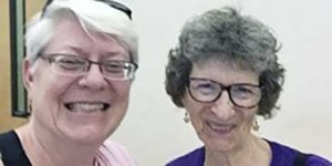 Two women, Beth Blissman and Jean East, smiling brightly for a picture together indoors in front of a plain white wall. One woman has short white hair and a pink shirt, the other woman has short dark grey hair and a dark purple shirt, both of them are wearing reading glasses.