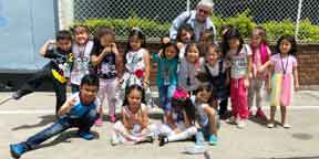 A woman smiling brightly with a large group of young children outdoors.