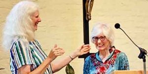 Two women with white hair smiling while one applauds while talking into a microphone on a podium at an indoor venue with white brick walls.