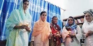 Five women wearing headscarves standing together in a line holding candles while looking towards the camera. They are standing in a room with white walls, a large dark book shelf, and bright blue curtains covering a window.