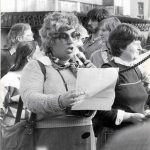 A black and white picture of a large crowd of people focusing on one woman in particular with large round glasses holding a piece of paper and microphone while speaking passionately.