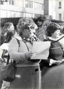 A black and white picture of a large crowd of people focusing on one woman in particular with large round glasses holding a piece of paper and microphone while speaking passionately.