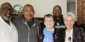 A group picture indoors of three tall men with dark skin complexions, and two shorter women with lighter skin complexions.