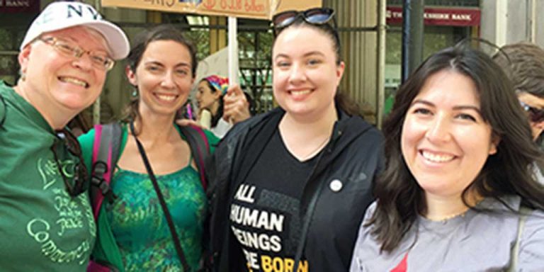 Four women smiling together for a group selfie on a sunny day outdoors. Two of them are wearing matching green graphic t-shirts.