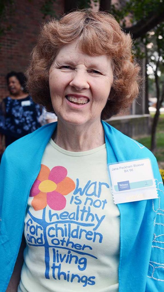 A woman, Jane Peckham, with short strawberry blonde hair smiling brightly for a picture outdoors at a social event wearing a blue cardigan, graphic t-shirt, and a name tag.