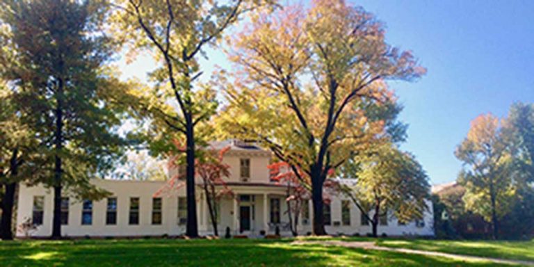 A large white house hidden in a tree line of big aspen and oak trees. It is a sunny autumn day, the leaves start changing colors to yellow and orange.