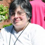 A woman, Mary Gutzwiller, with short dark grey hair, and wire-rimmed glasses wearing a white t-shirt and black lanyard smiling for a picture outdoors on a sunny day in front of a brick building with a well-manicured lawn and bushes.